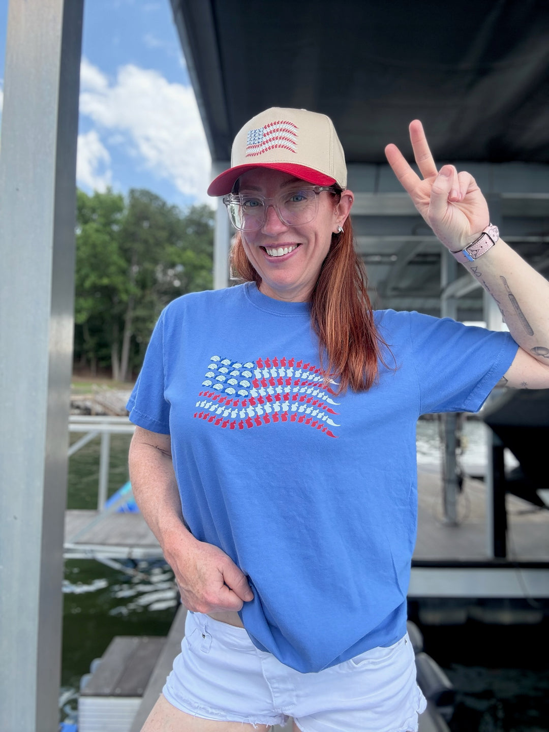 Ladyspinedoc wearing the Stars, Stripes & Synapses tee flashing a peace sign on a dock, with hat matching the neuro flag design.