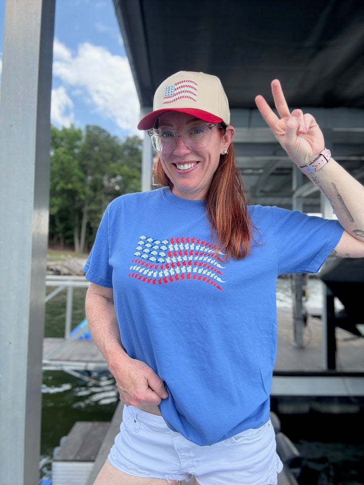 Ladyspinedoc wearing the Stars, Stripes & Synapses tee flashing a peace sign on a dock, with hat matching the neuro flag design.