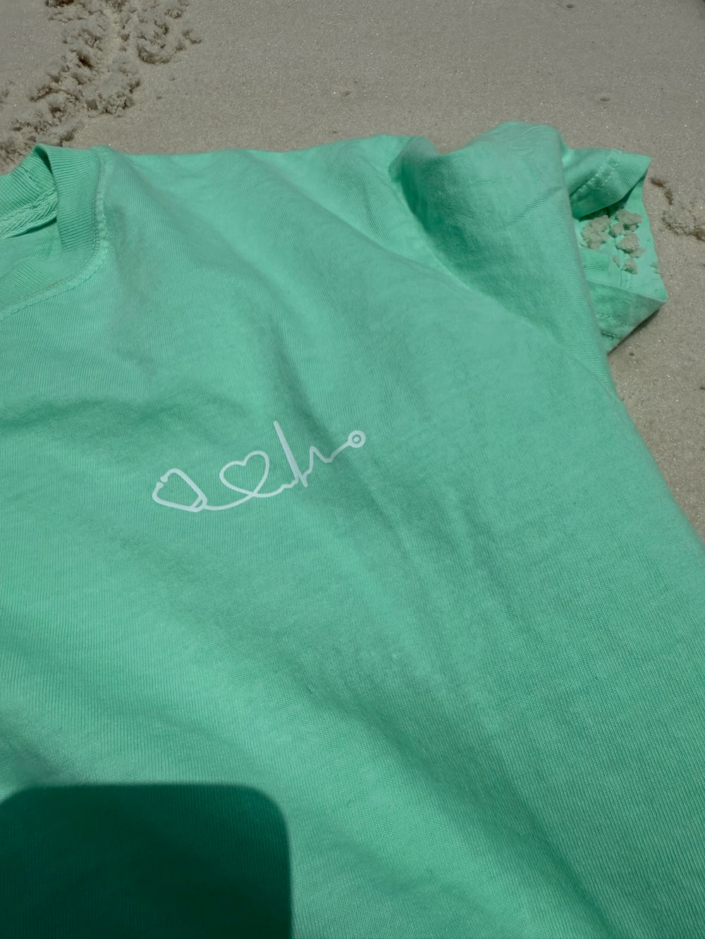 Close up of light green Heart of Medicine t-shirt featuring Dr. Beachgem’s stethoscope and heart waveform logo on the upper left chest, displayed on sandy beach.