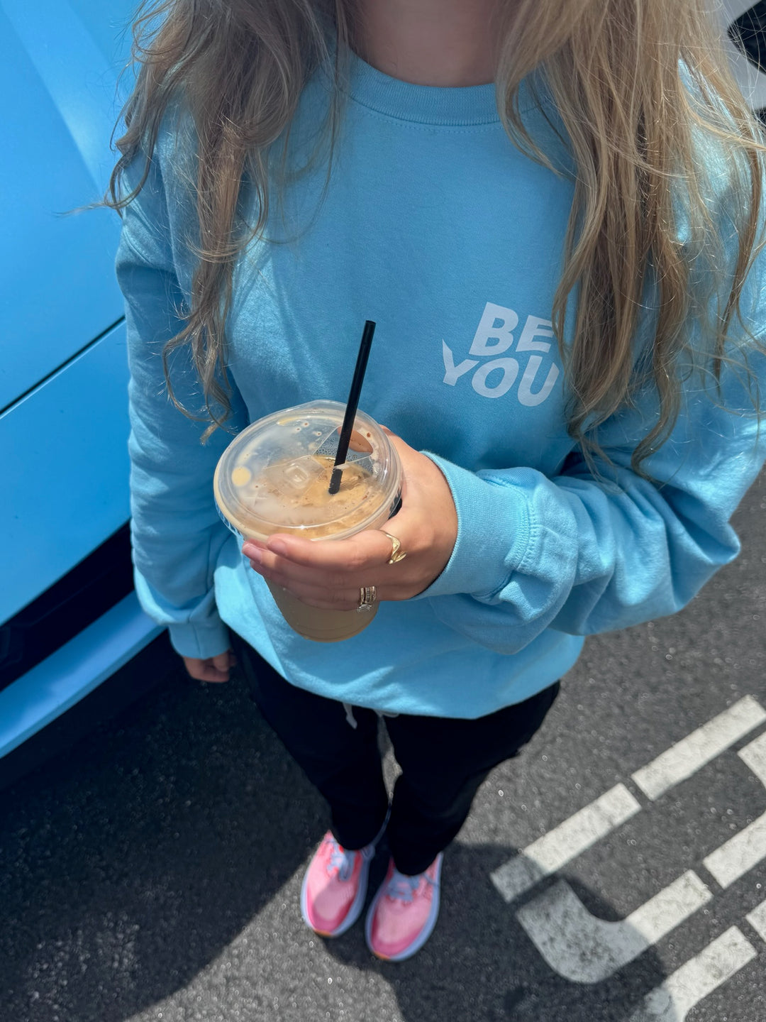 Woman holding iced coffee and wearing BE YOU Crewneck Sweatshirt in Light Blue