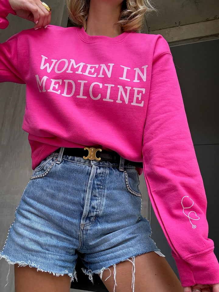 Model wearing hot pink Women in Medicine sweatshirt with heart shaped stethoscope design on left sleeve.