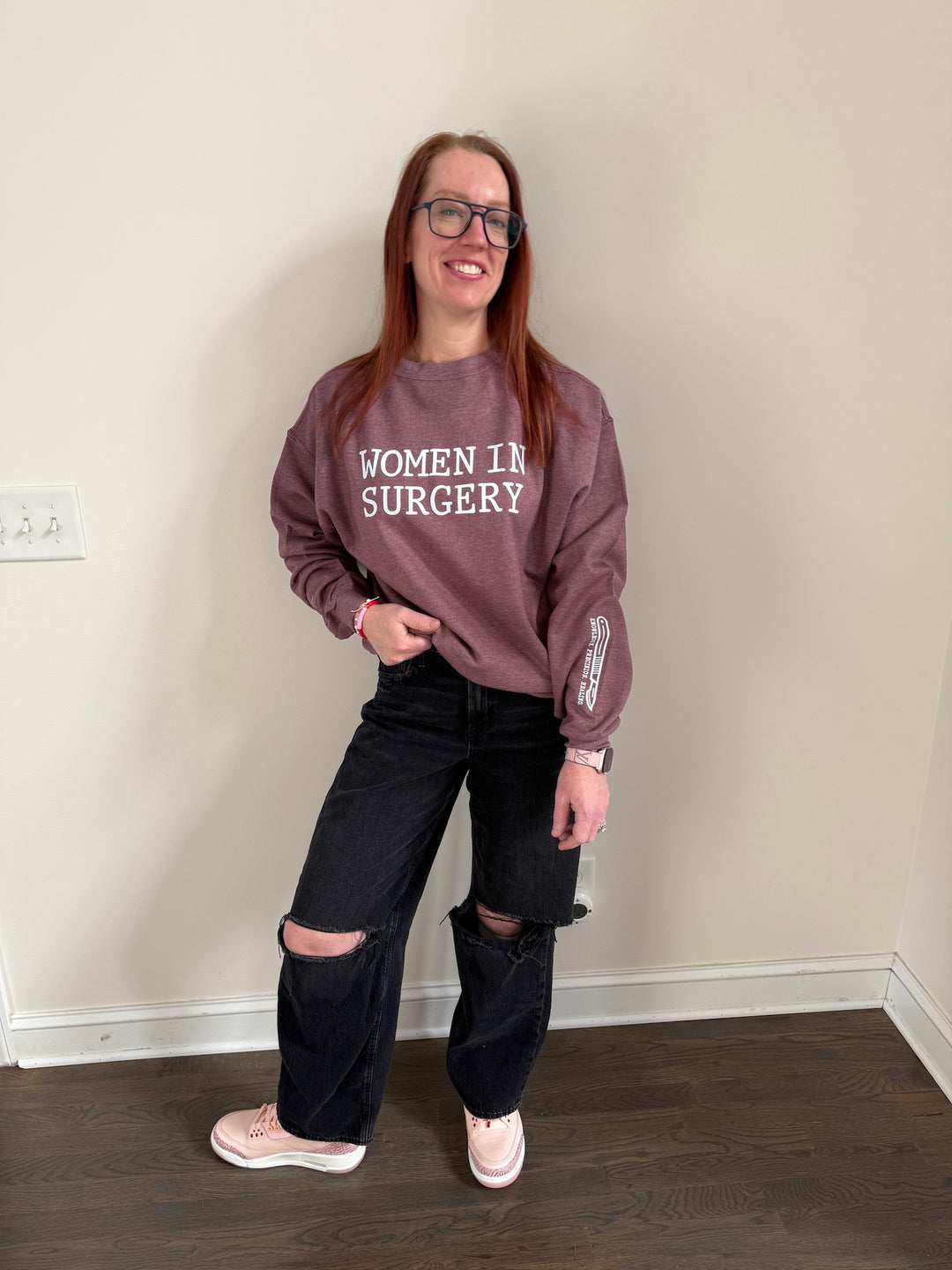 Dr. Betsy Grunch - Ladyspinedoc wearing a heather maroon crewneck sweatshirt with "WOMEN IN SURGERY" printed in bold white letters across the chest and a white scalpel design with the words "KNOWLEDGE. PRECISION. HEALING." printed on the bottom left sleeve.