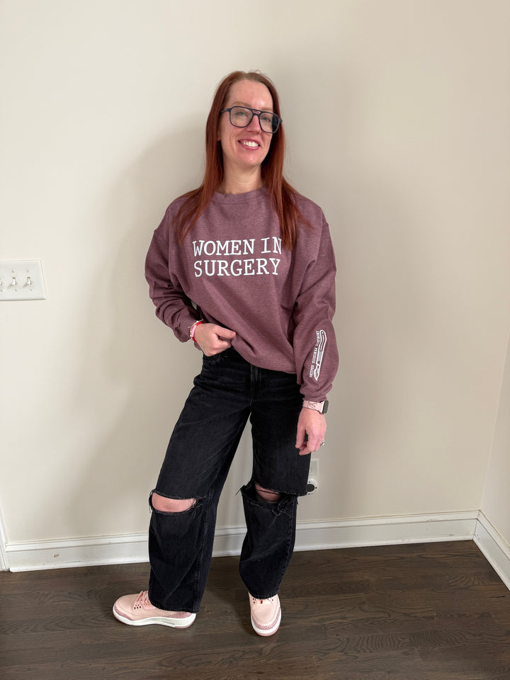 Dr. Betsy Grunch - Ladyspinedoc wearing a heather maroon crewneck sweatshirt with "WOMEN IN SURGERY" printed in bold white letters across the chest and a white scalpel design with the words "KNOWLEDGE. PRECISION. HEALING." printed on the bottom left sleeve.