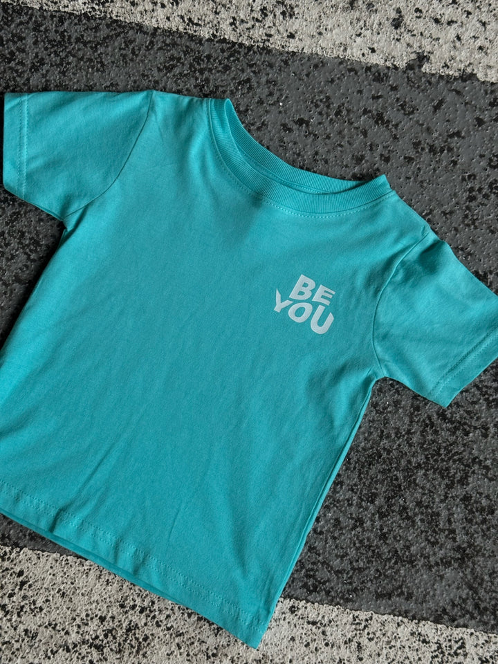 Flat lay of light blue toddler t-shirt with “BE YOU” on front left chest displayed on pavement.
