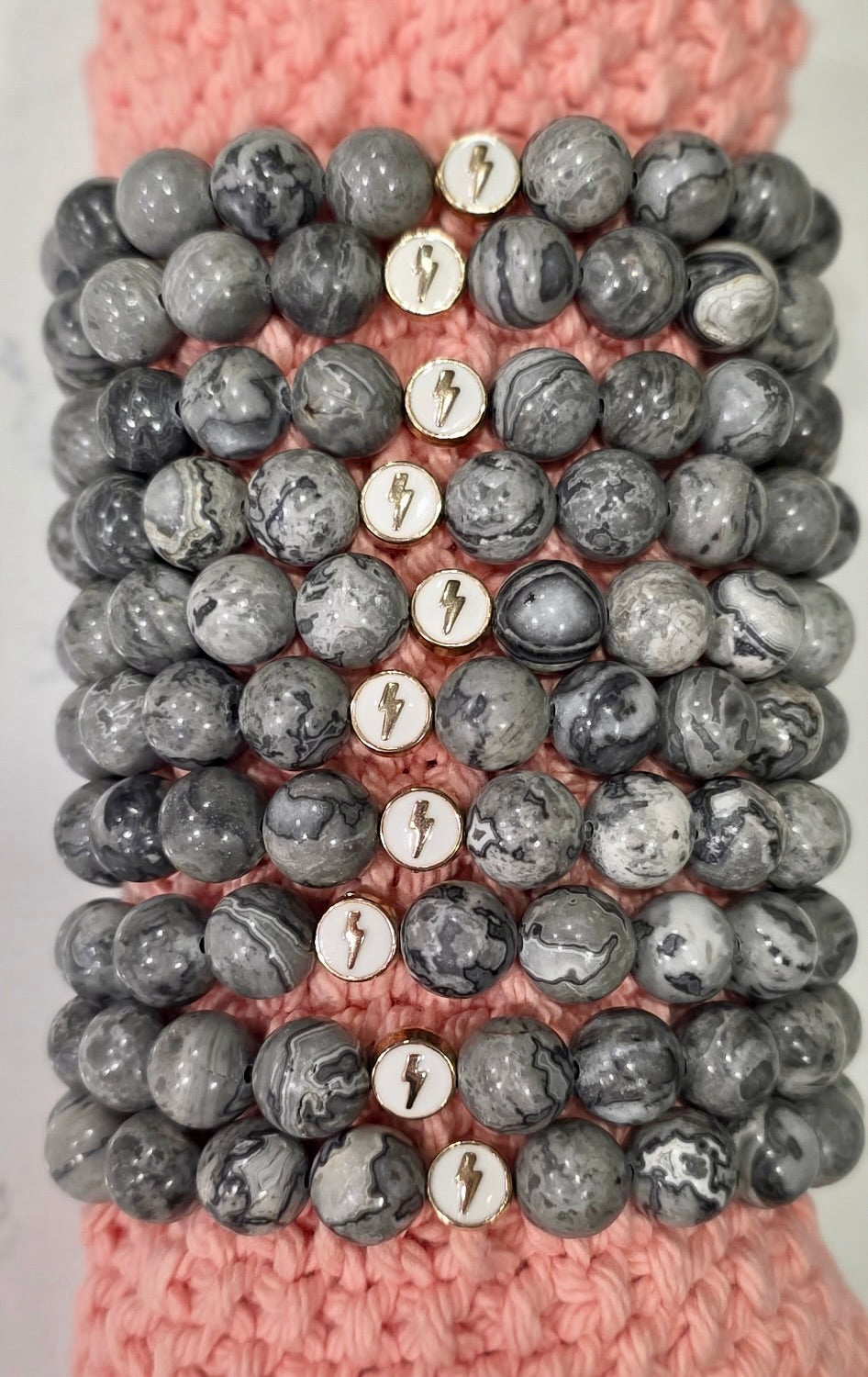 Close-up of stacked Map Stone Jasper glioblastoma awareness bracelets with white enamel lightning bolt charms, shown on a pink knit background.
