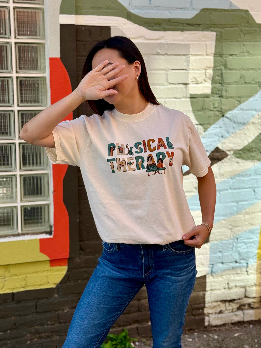 Woman wearing an ivory 'Physical Therapy' t-shirt in front of a colorful mural.