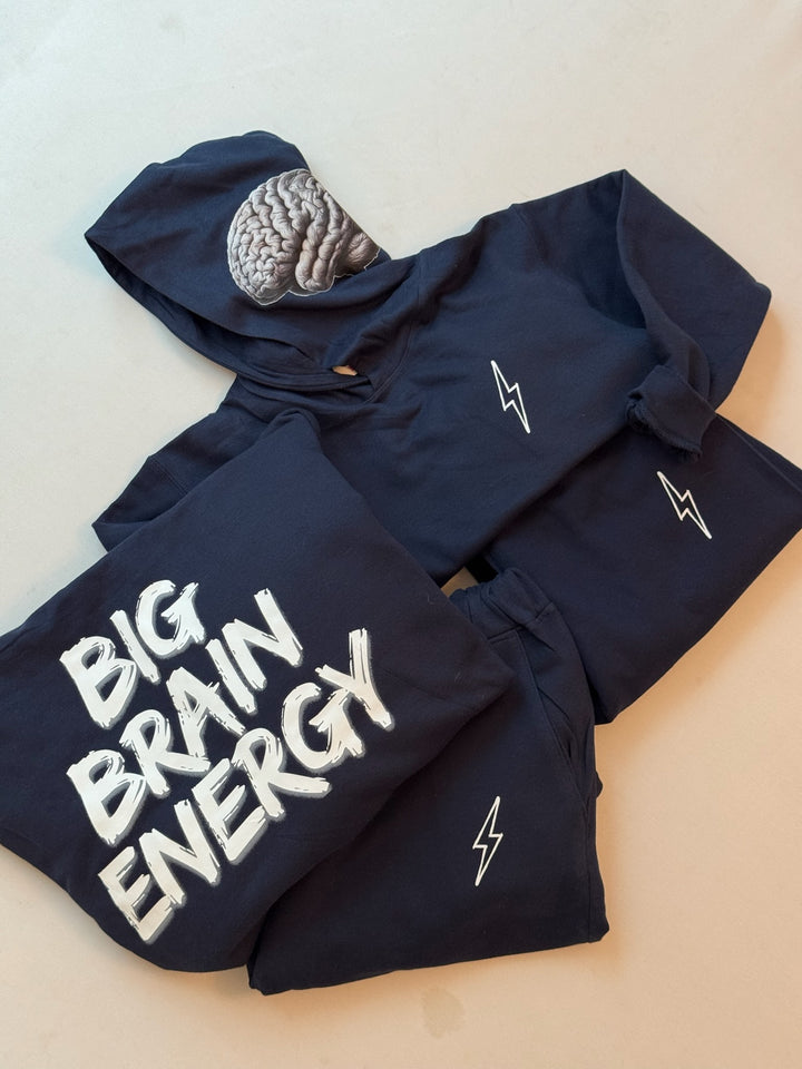 Adult and youth folded BIG BRAIN ENERGY hoodies on top of folded matching sweatpants. One sweatshirt shows the front with lightning bolt on chest and the brain design on hood. The other folded sweatshirt shows the back with the BIG BRAIN ENERGY print. The sweatpants show the lightning bolt logos.