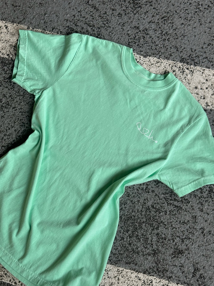Flat lay of light green (Island Reef) Heart of Medicine t-shirt featuring Dr. Beachgem’s stethoscope and heart waveform logo on the upper left chest.