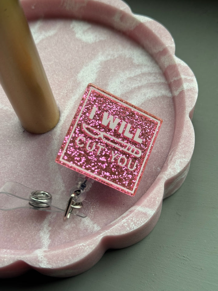 Pink glitter square badge reel with the phrase “I Will Cut You” printed in white, featuring a clear retractable clip for ID or name badge attachment, showcased in pink tray.
