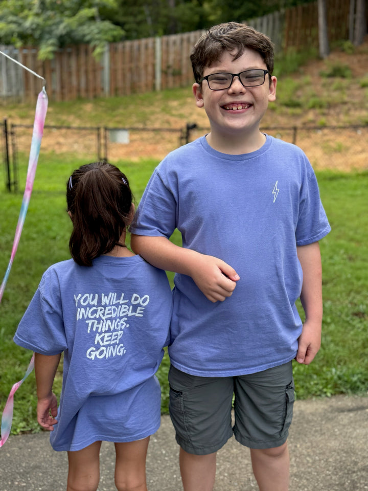 Girl and boy each wearing the violet  Incredible Youth Tee, hers showing the back message, "You will do incredible things. Keep Going." and his showing the front with the lightning bolt on upper left chest.