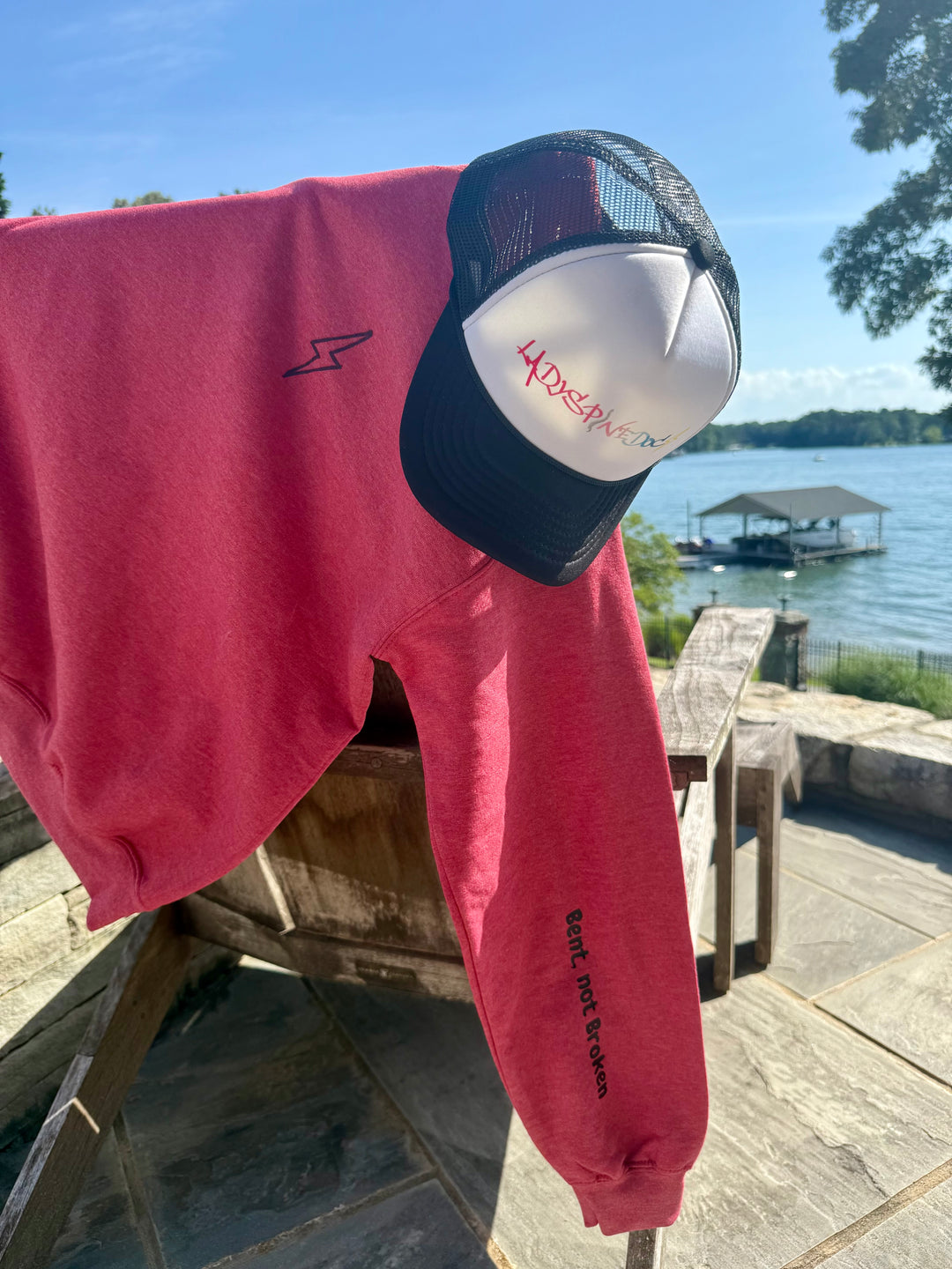 Bent Not Broken Scoliosis Crewneck Sweatshirt – Heather Red hanging on the back of a chair, showing the lightning bolt on the front and the Bent, not Broken catchphrase on the sleeve. Ladyspinedoc trucker hat also displayed.