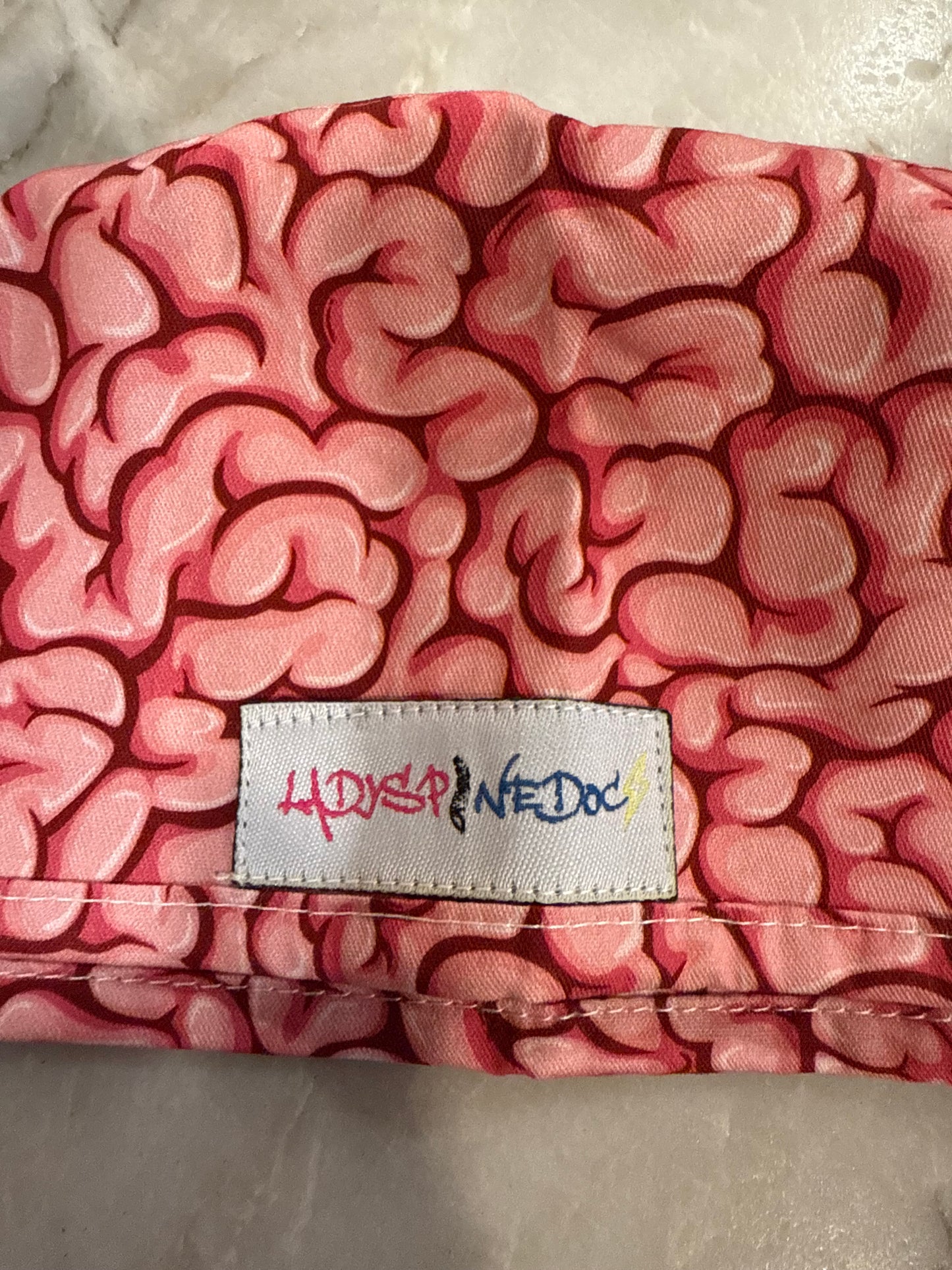 Close up of the brain print and Ladyspinedoc logo tag sewn on the outer side of the pink Cortex Scrub Hat