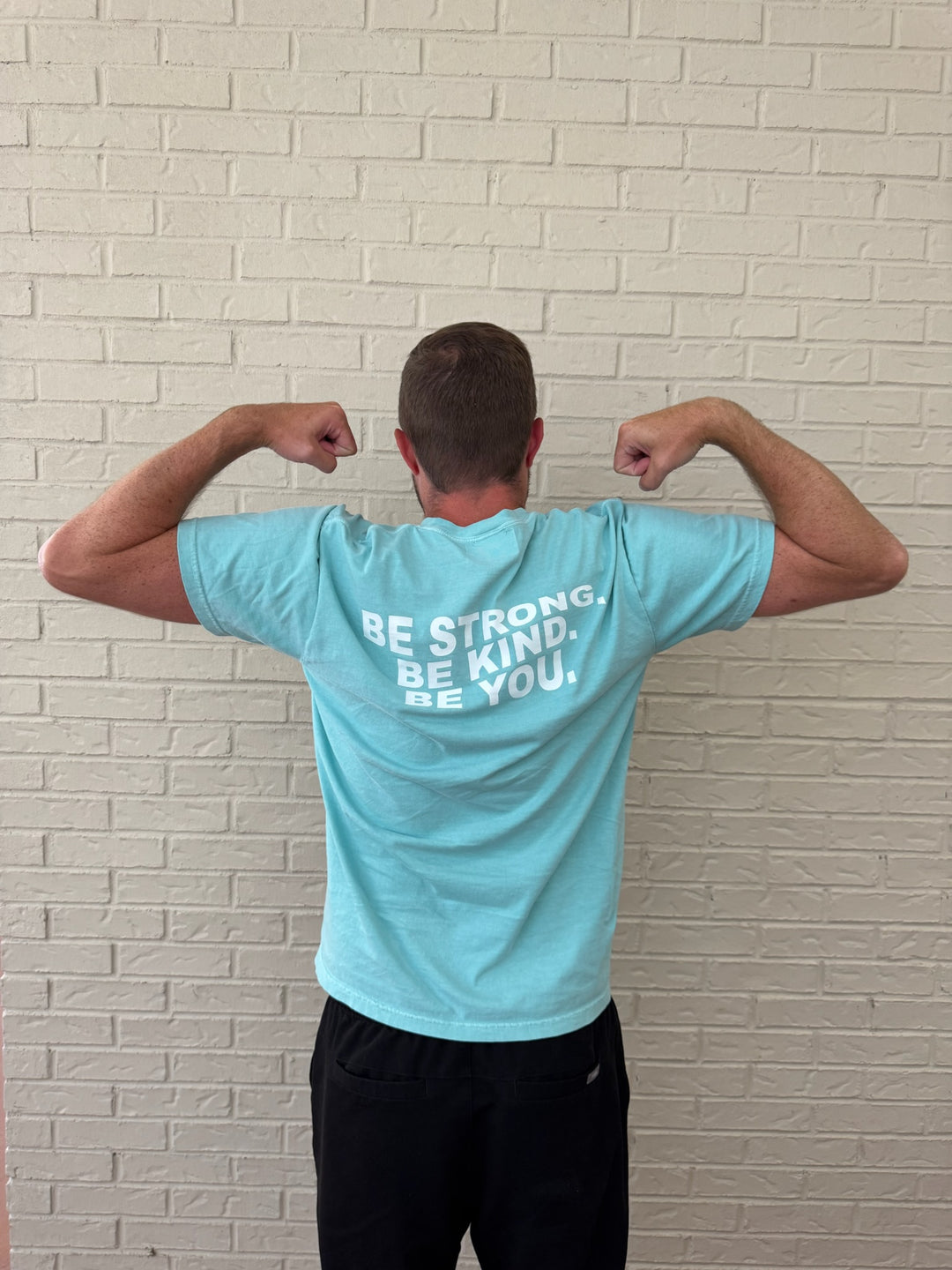Back of male model doing a arm muscle stance and wearing the BE YOU Tee Shirt, showing the BE STRONG. BE KIND. BE YOU. motto on the back.