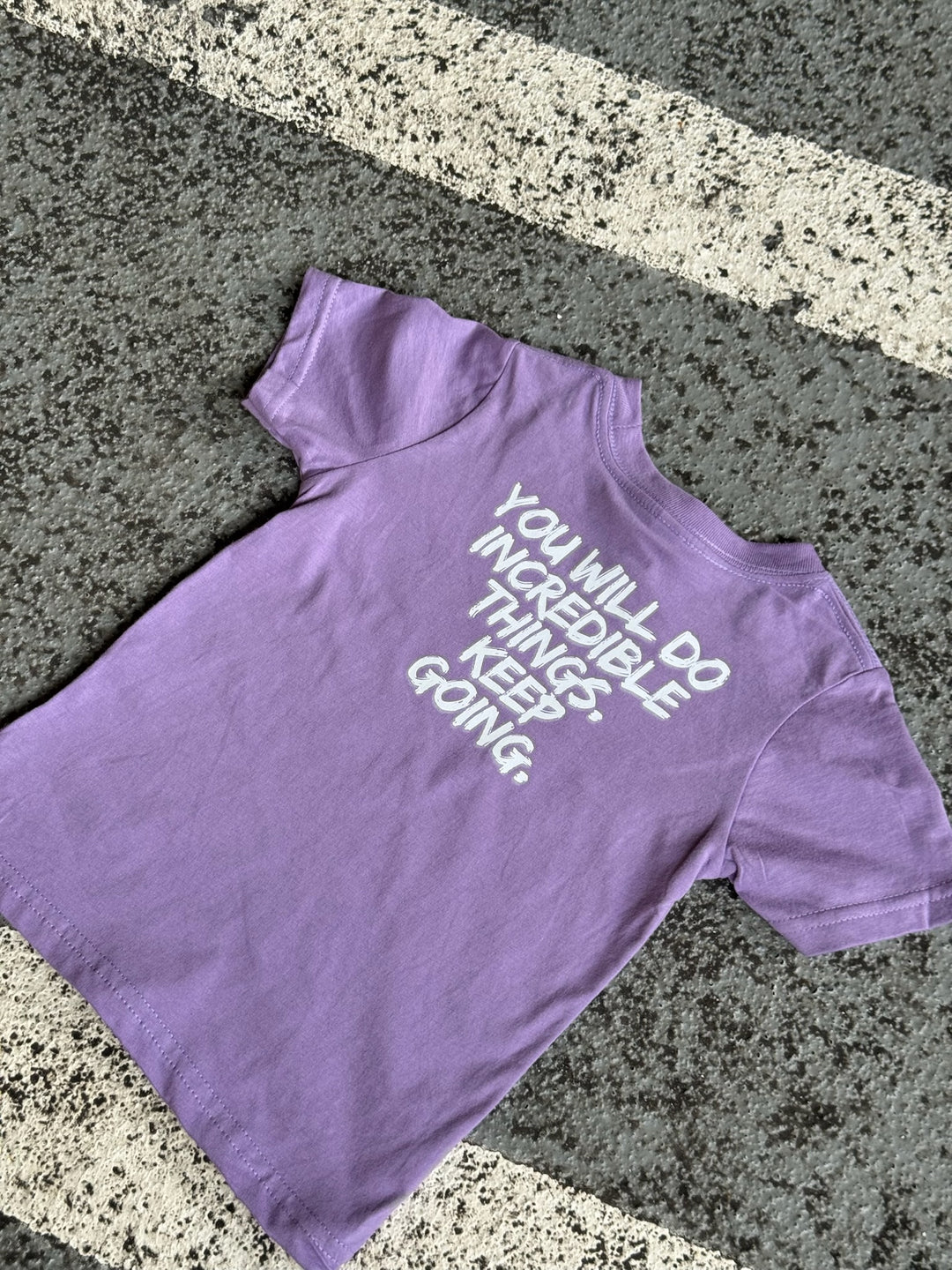 Flat lay of back of purple toddler tee with bold white brushstroke quote: “YOU WILL DO INCREDIBLE THINGS. KEEP GOING.” displayed on pavement.
