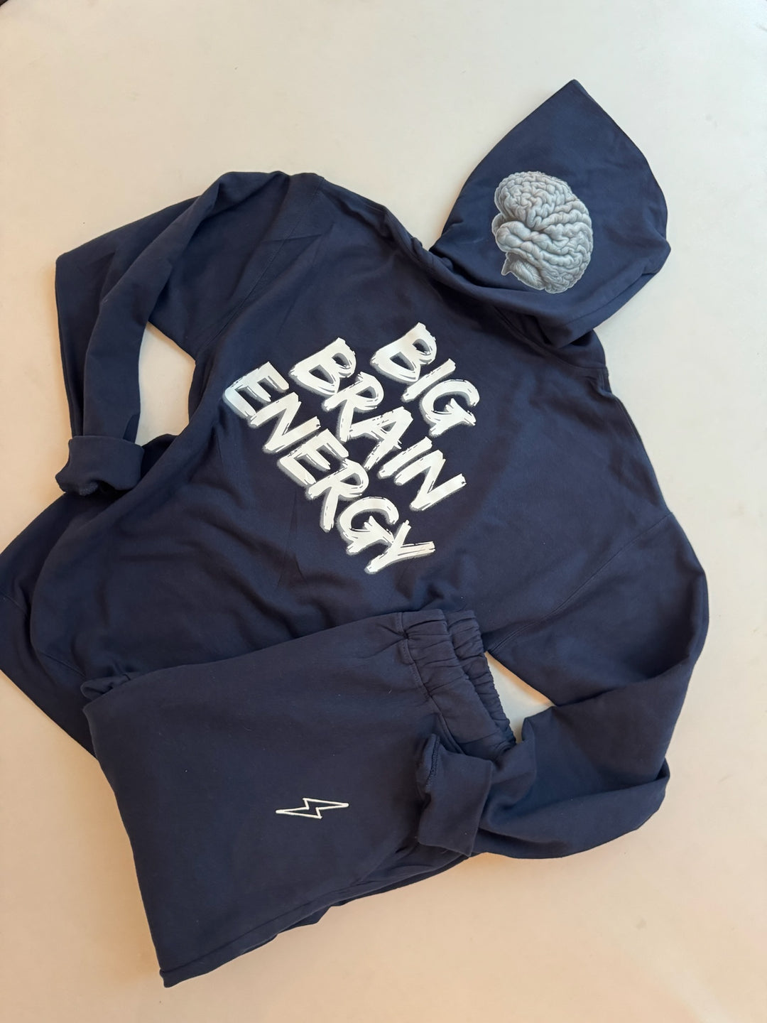 Back flat lay of youth navy hoodie showing the BIG BRAIN ENERGY print on the back and brain design on left side of hood. Folded youth BIG BRAIN ENERGY sweatpants with lightning bolt logo also shown.