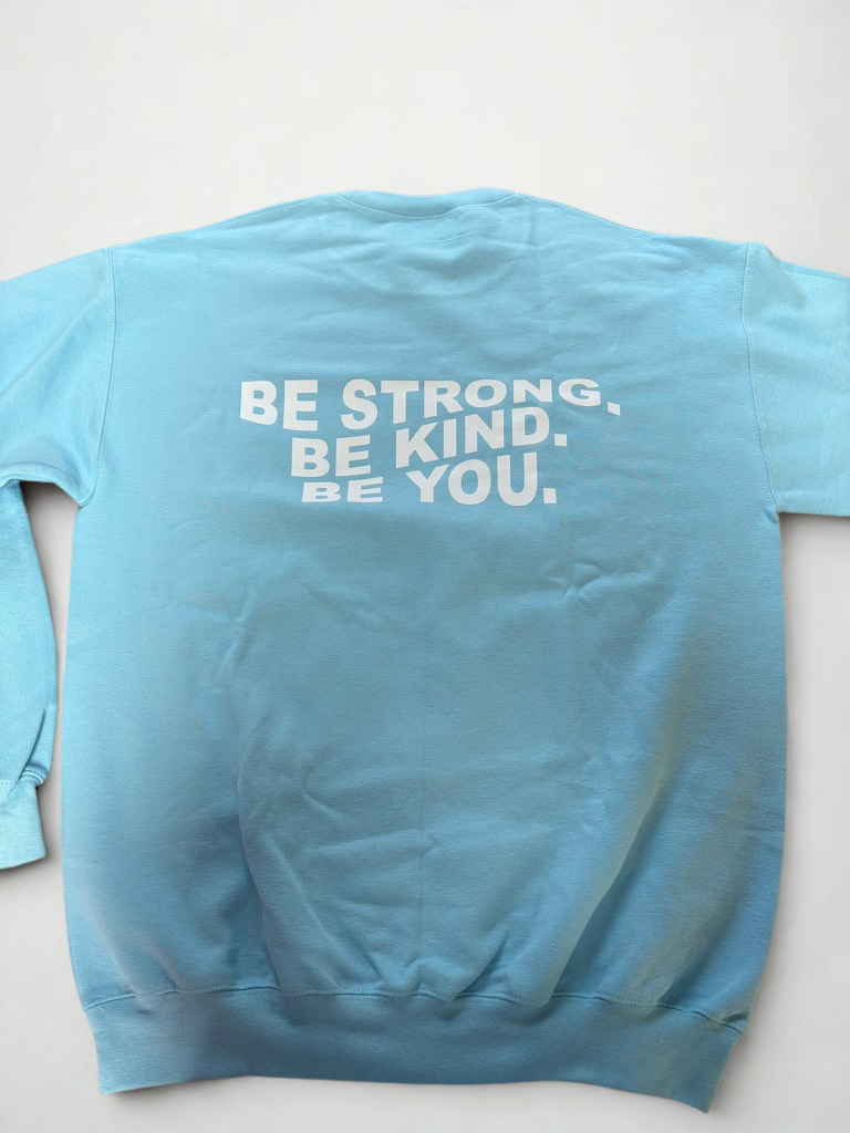 Light blue sweatshirt showing large text print: "Be Strong. Be Kind. Be You." across the upper back.