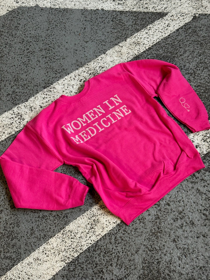 Flat lay of hot pink Women in Medicine Sweatshirt showing heart stethoscope design on left sleeve