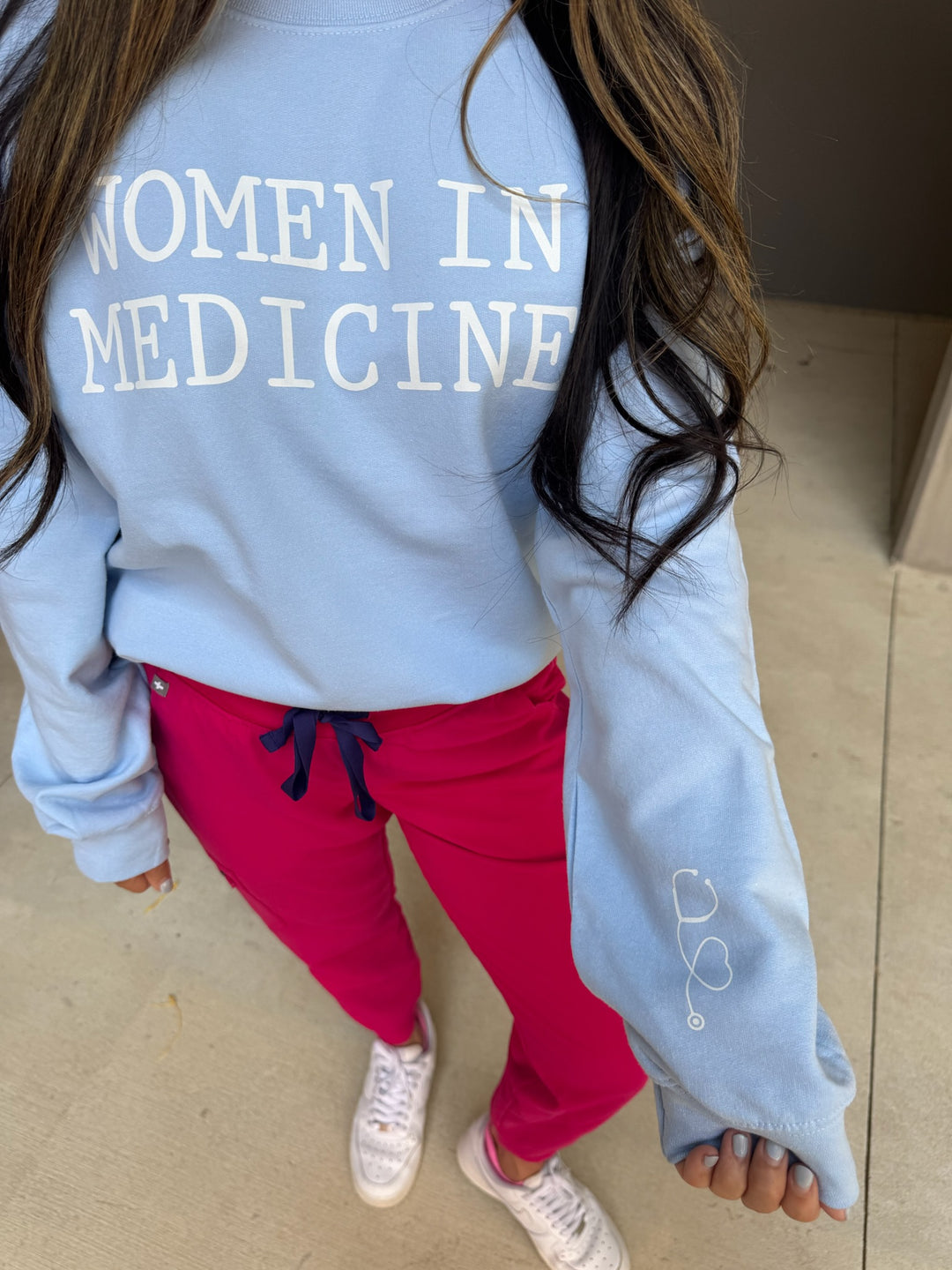 Model wearing light blue Women in Medicine sweatshirt with heart shaped stethoscope design on left sleeve.