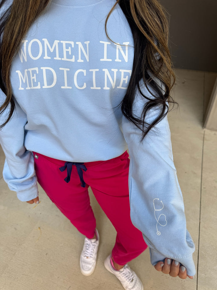 Model wearing light blue Women in Medicine sweatshirt with heart shaped stethoscope design on left sleeve.