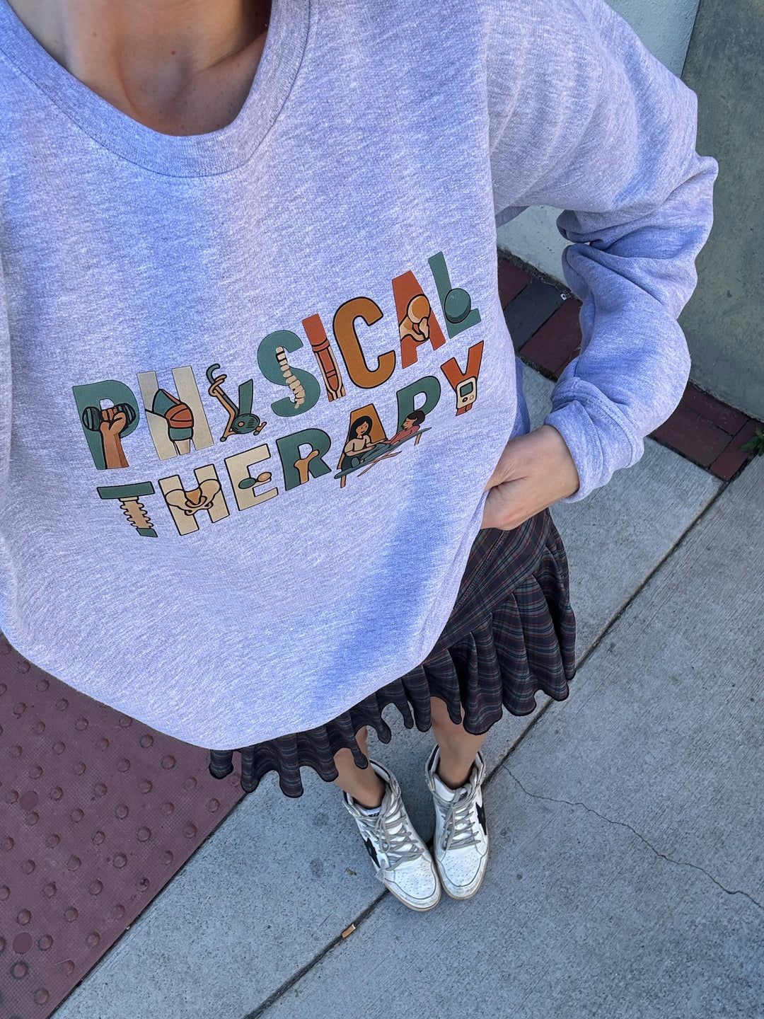 Person wearing a gray sweatshirt with colorful 'Physical Therapy' text on a sidewalk.