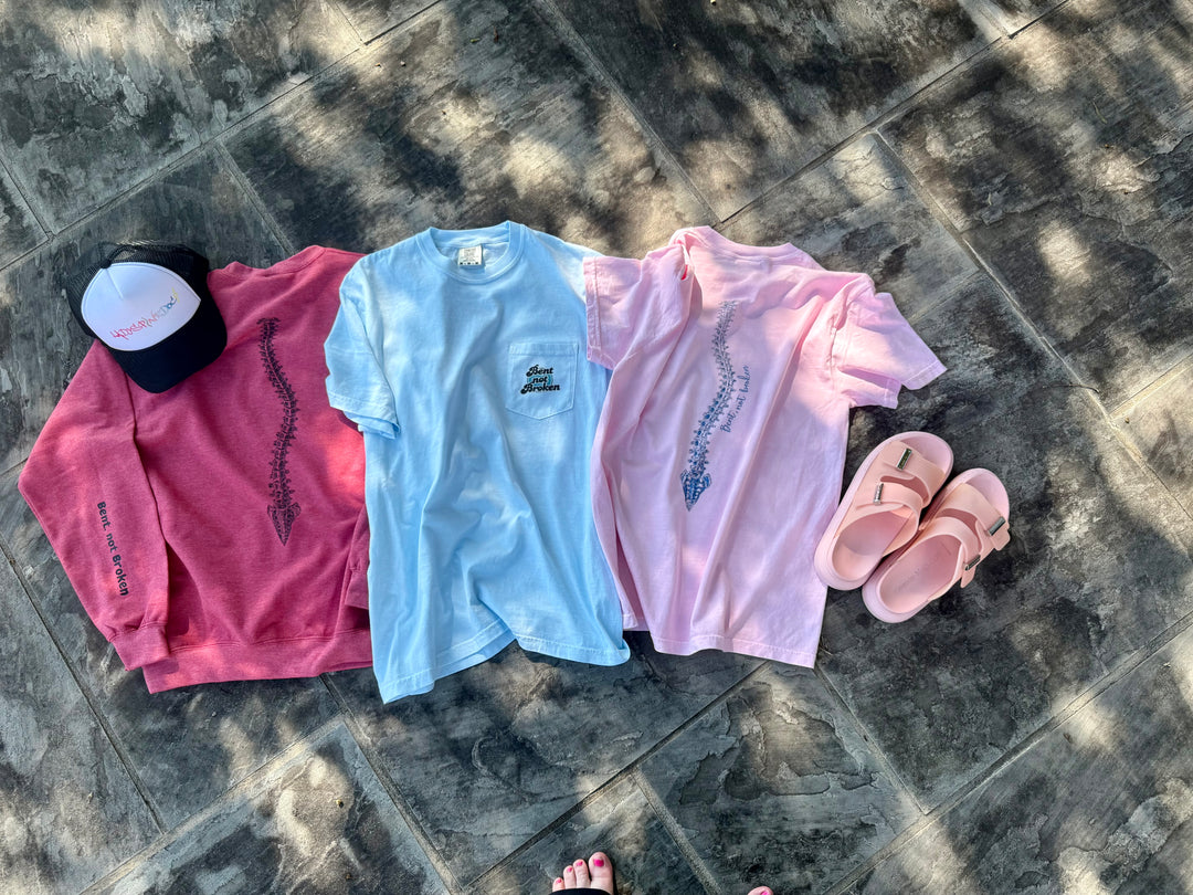 Flat lays of the three scoliosis awareness shirts in the Ladyspinedoc collection, two t-shirts and one sweatshirt, displayed with Ladyspinedoc trucker hat and sandals.