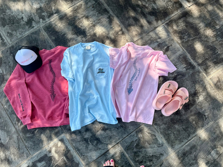 Flat lays of the three scoliosis awareness shirts in the Ladyspinedoc collection, two t-shirts and one sweatshirt, displayed with Ladyspinedoc trucker hat and sandals.