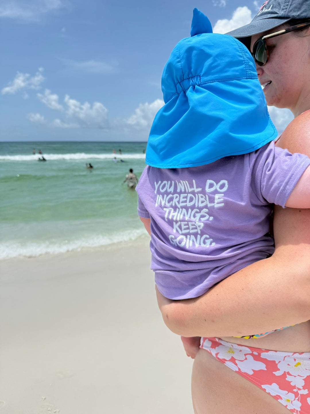 Mom at beach holding Toddler who is wearing the Incredible Tee with bold white brushstroke quote: “YOU WILL DO INCREDIBLE THINGS. KEEP GOING.” shown on the back.