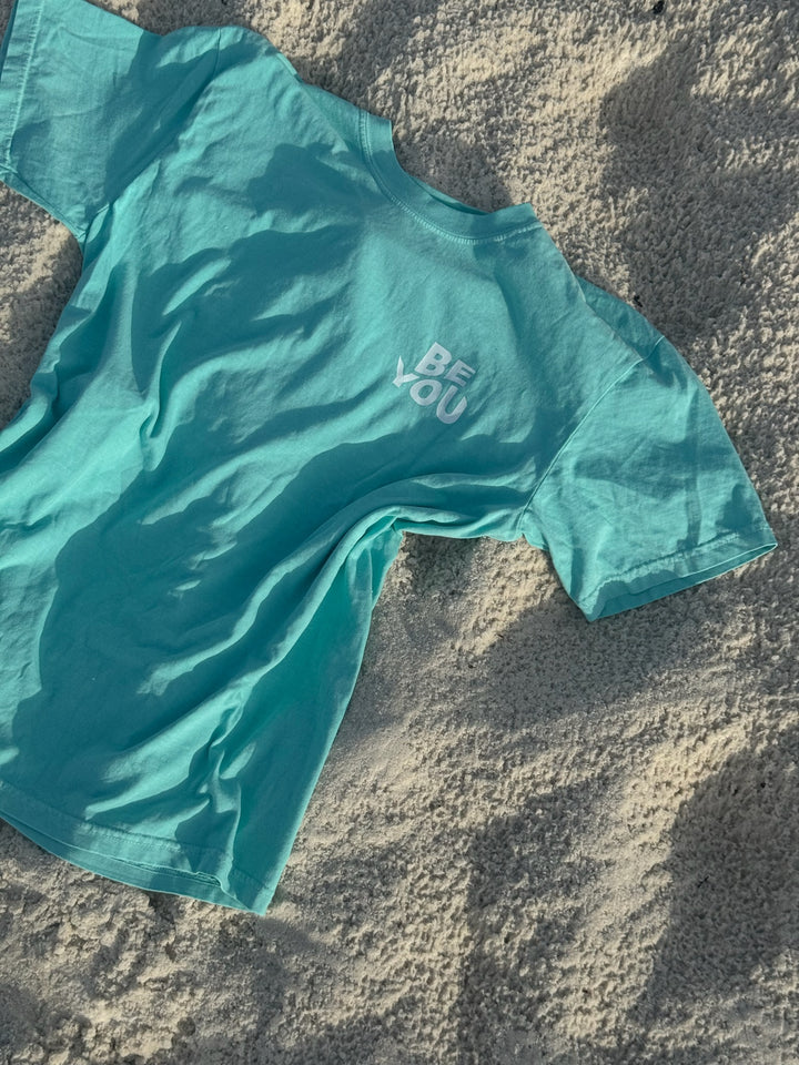Front flat lay of BE YOU Tee Shirt – BE STRONG. BE KIND. BE YOU. in teal laying on sand.