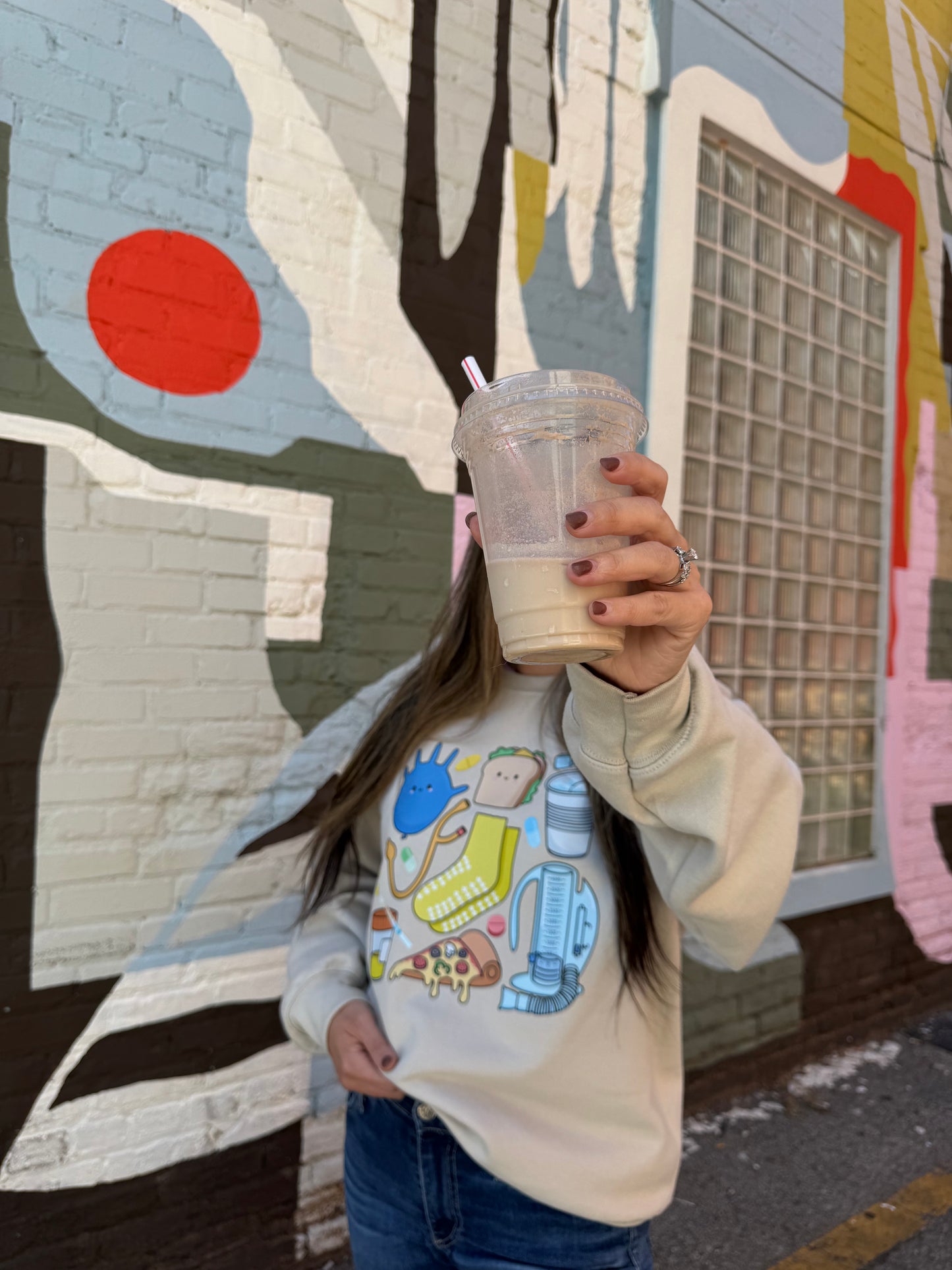 Women hiding her face with a cup and wearing a beige Nurse Sweatshirt with cartoon nurse memorabilia on the front.
