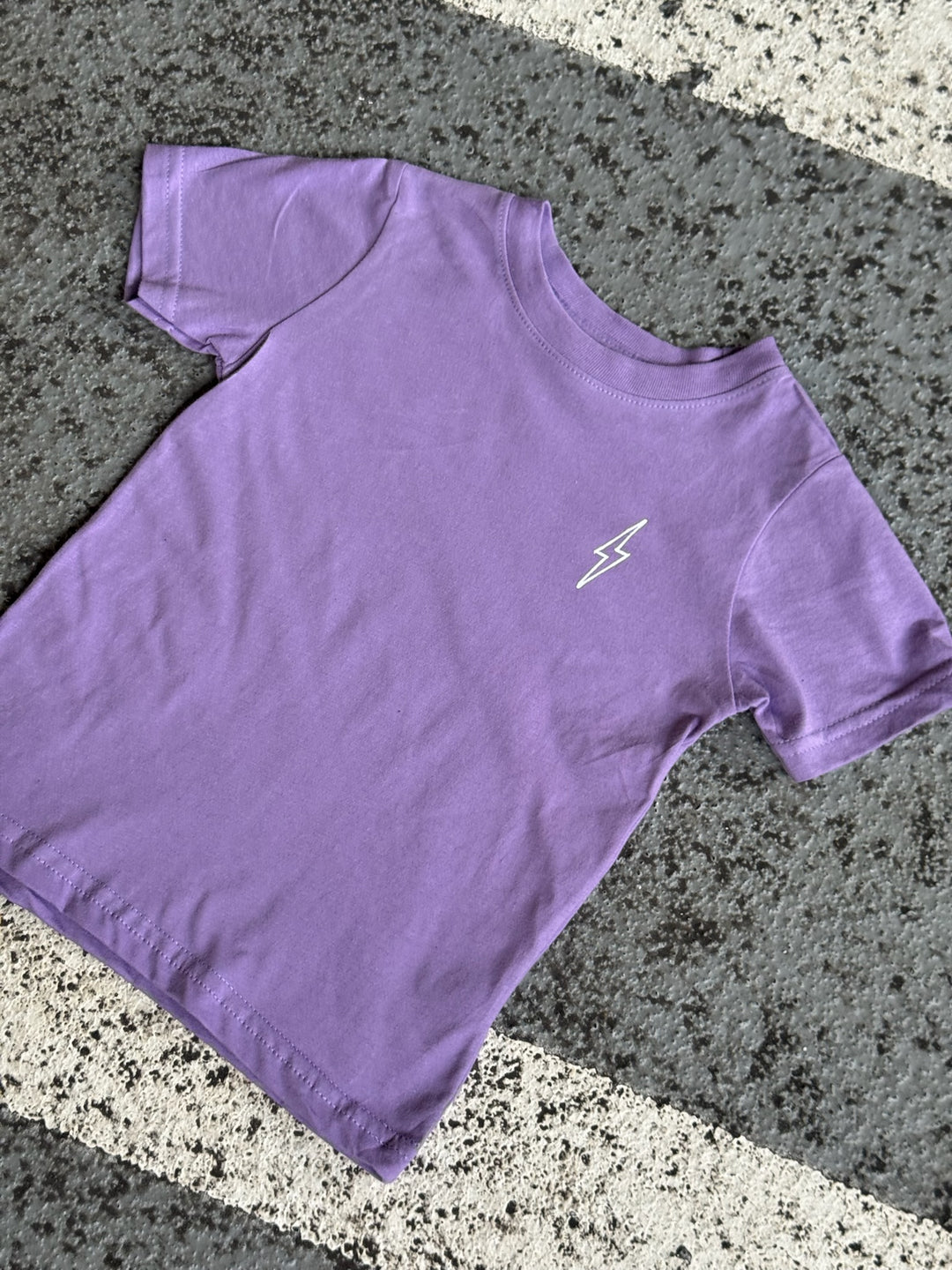 Flat lay of purple toddler tee with Ladyspinedoc lightning bolt on left chest on pavement.