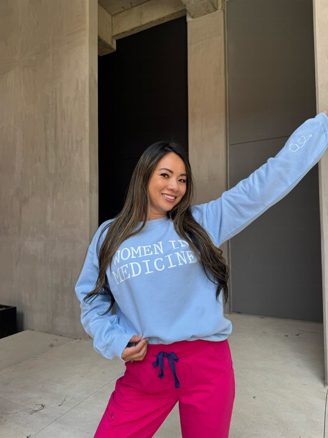 Smiling model with her arm in the air wearing light blue Women in Medicine sweatshirt with heart shaped stethoscope design on left sleeve.