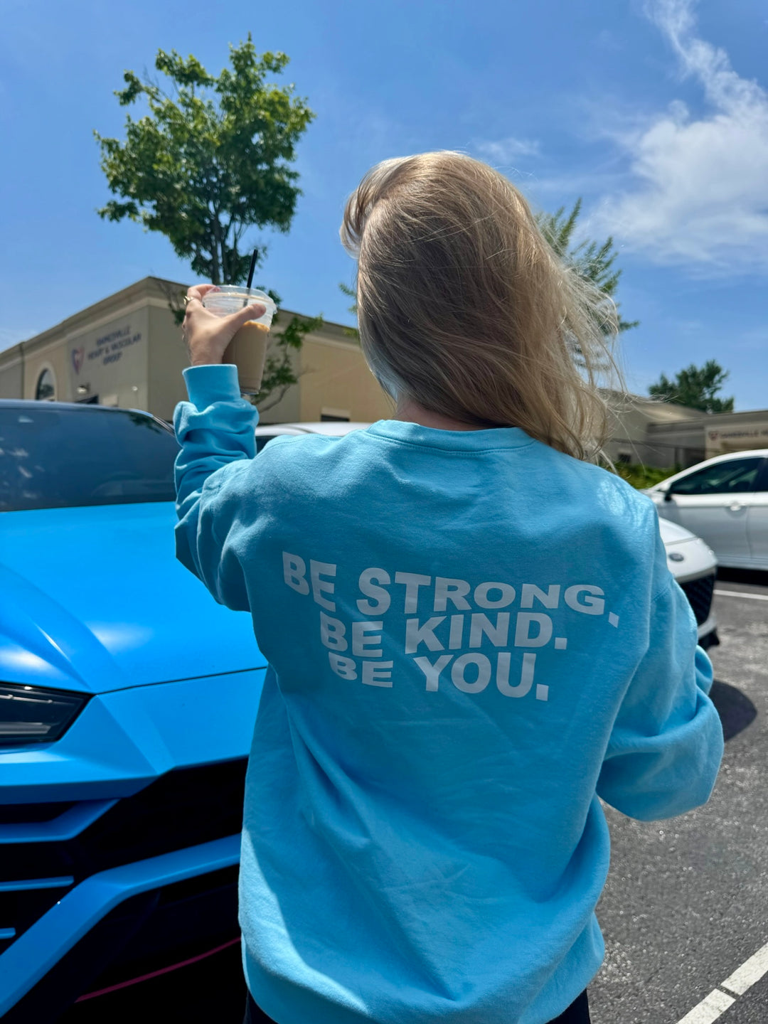 Woman wearing a light blue crewneck sweatshirt with “BE STRONG. BE KIND. BE YOU.” printed on the back in bold white letters.