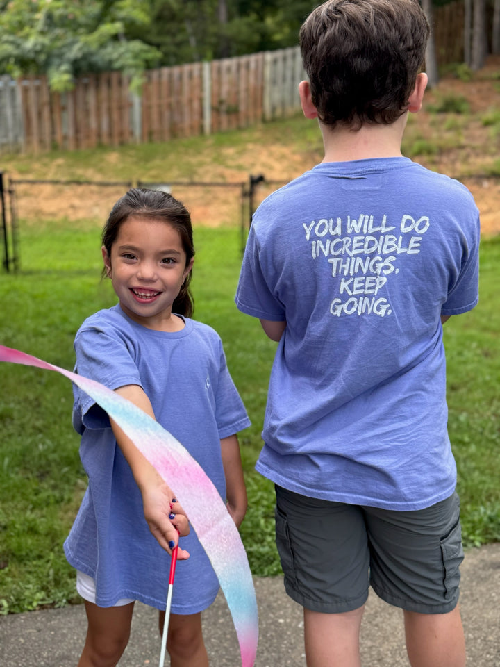Girl and boy each wearing the violet  Incredible Youth Tee, his showing the back message, "You will do incredible things. Keep Going." and her showing the front, while playing with a gymnastics ribbon.