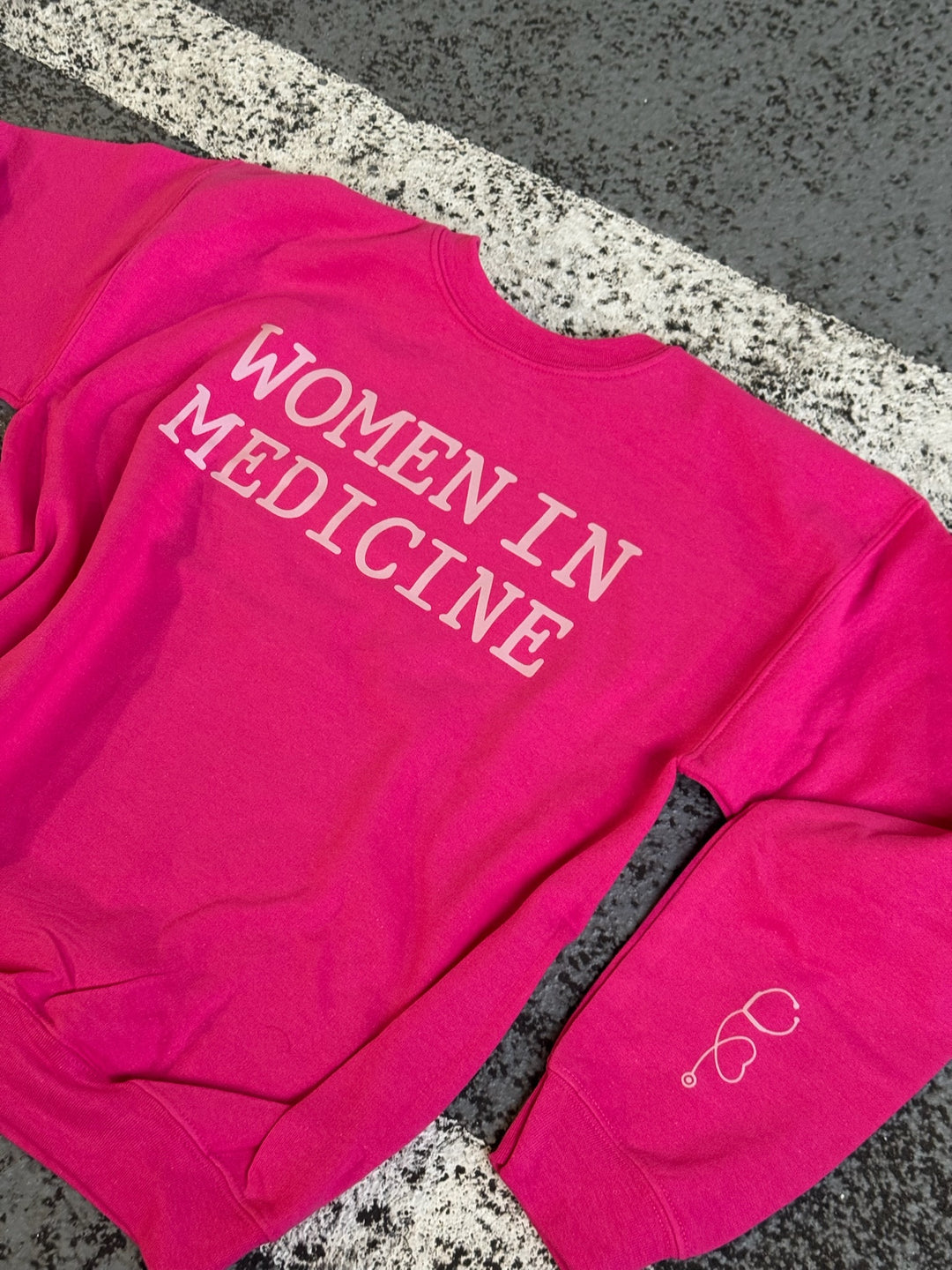 Flat lay of hot pink Women in Medicine Sweatshirt on pavement, showing left sleeve with stethoscope heart design