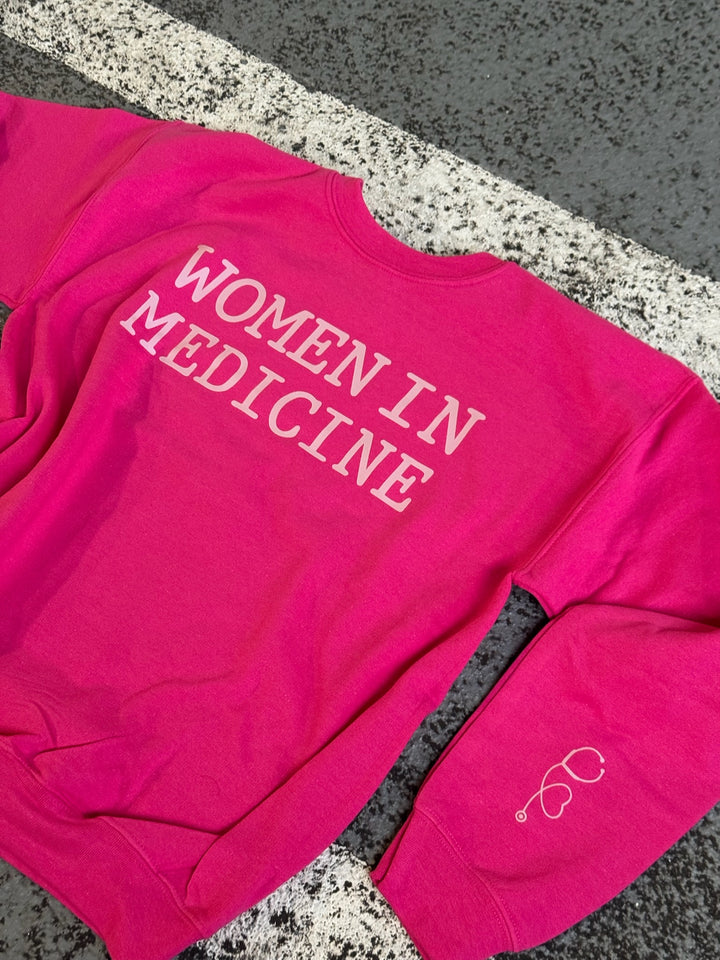 Flat lay of hot pink Women in Medicine Sweatshirt on pavement, showing left sleeve with stethoscope heart design