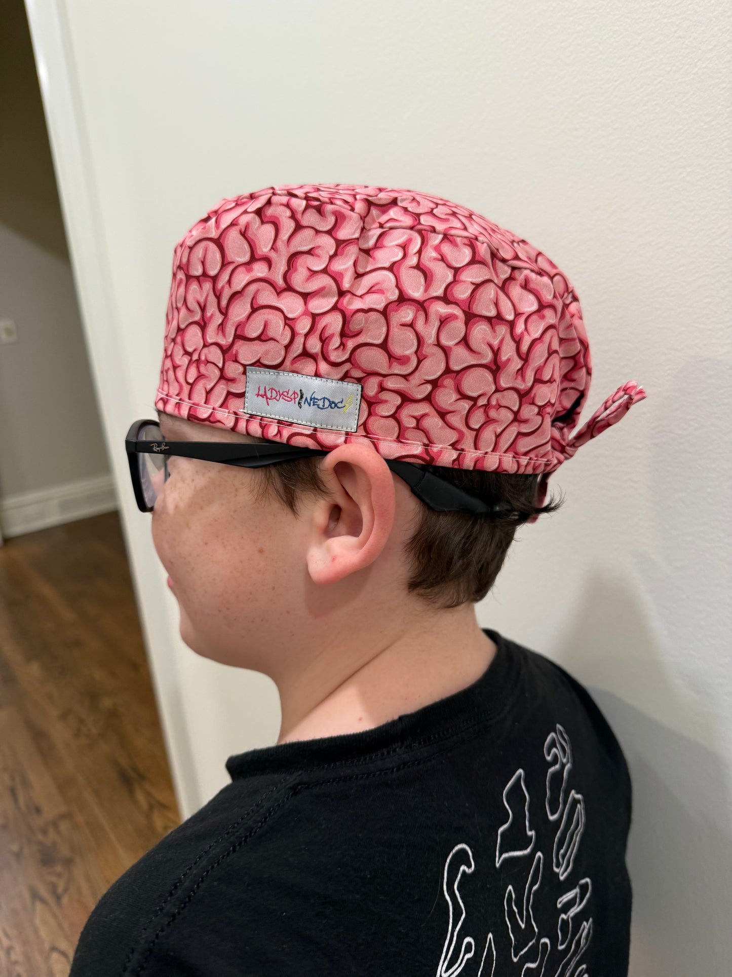 Side view of a youth wearing the tie-back (short hair) version of the Cortex Cap - Pink Scrub Hat with Brain Design showing the Ladyspinedoc logo tag sewn on the outer side