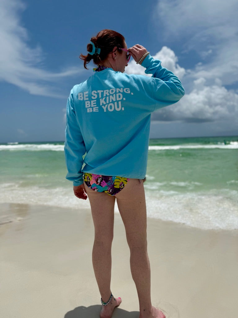 Ladyspinedoc wearing a light blue crewneck sweatshirt with “BE STRONG. BE KIND. BE YOU.” printed on the back in bold white letters, standing on a sunny beach — empowering beachwear for medical professionals and advocates.