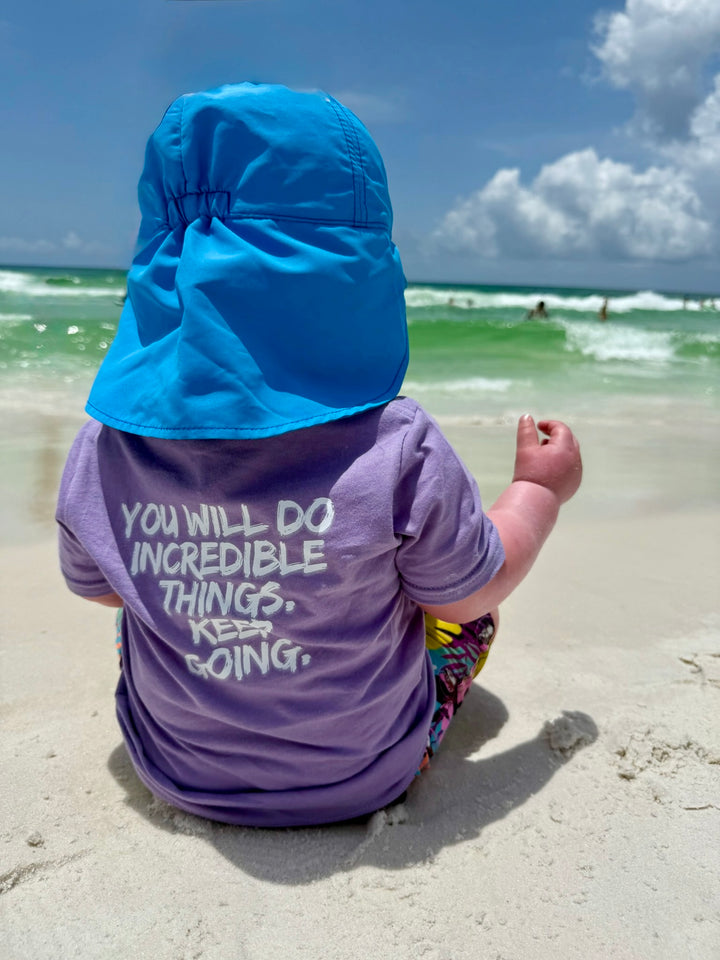 Toddler wearing the purple Incredible Toddler Tee at the beach showing the brushstroke quote: “YOU WILL DO INCREDIBLE THINGS. KEEP GOING.” on the back.