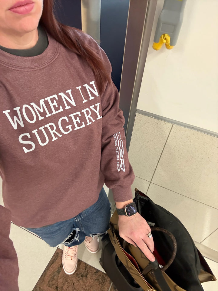 Dr. Betsy Grunch - Ladyspinedoc wearing a heather maroon crewneck sweatshirt with "WOMEN IN SURGERY" printed in bold white letters across the chest and a white scalpel design with the words "KNOWLEDGE. PRECISION. HEALING." printed on the bottom left sleeve.