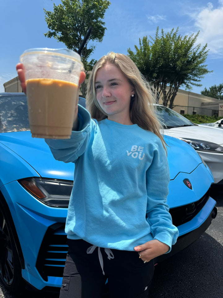 Woman holding up an iced coffee and wearing BE YOU Crewneck Sweatshirt in Light Blue