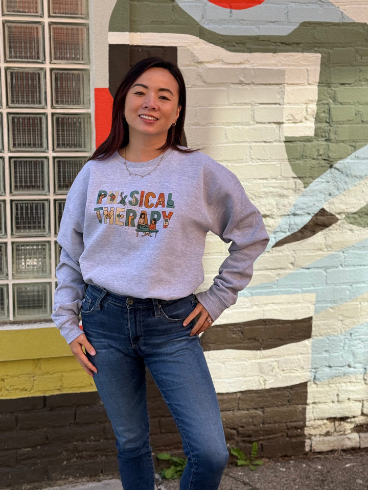 Person wearing a gray sweatshirt with 'Physical Therapy' text in colorful letters, standing in front of a mural.