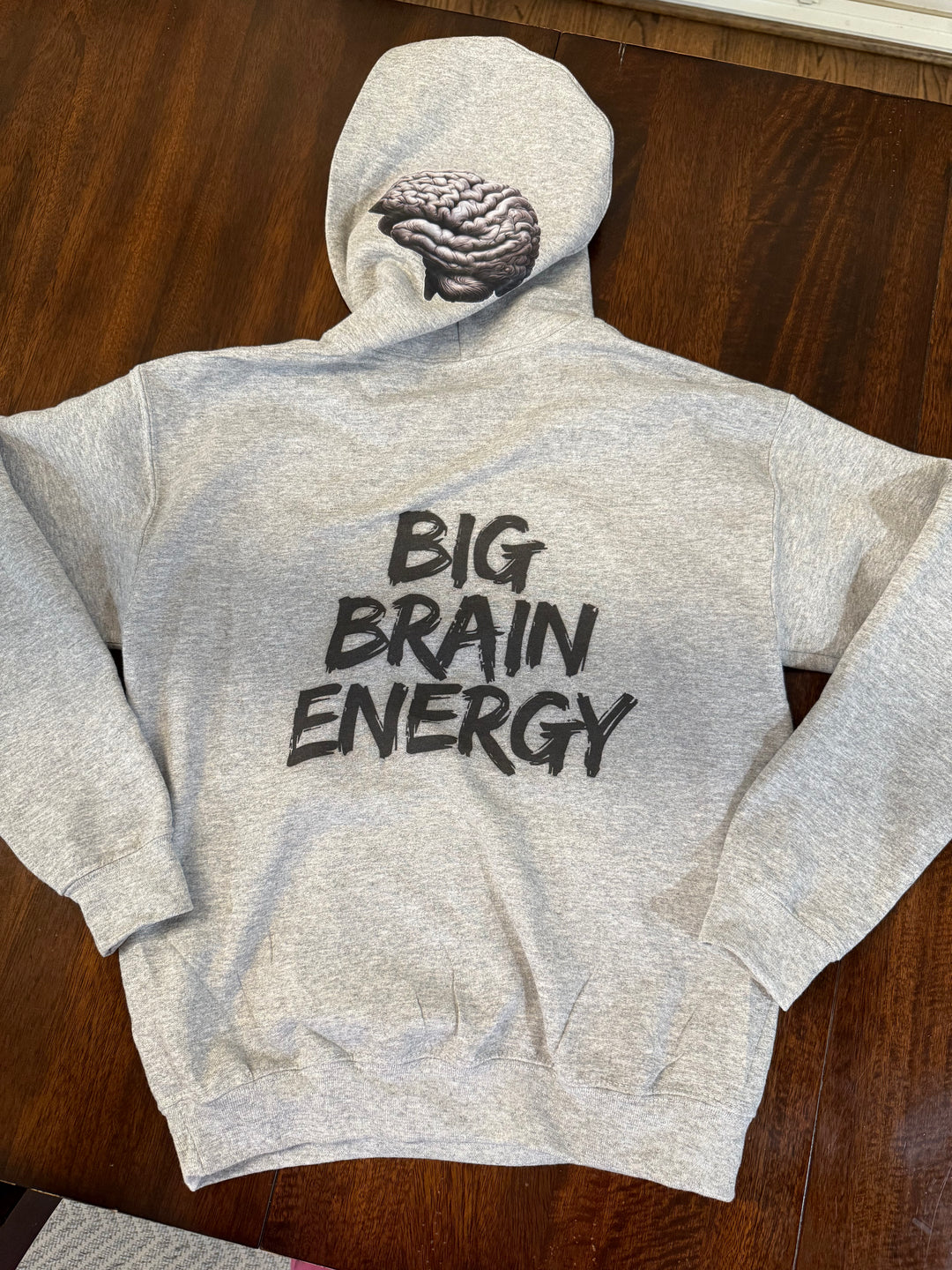 Light gray hoodie with "BIG BRAIN ENERGY" in bold black capital letters on the back and showing the brain design printed on the left side of the hood.