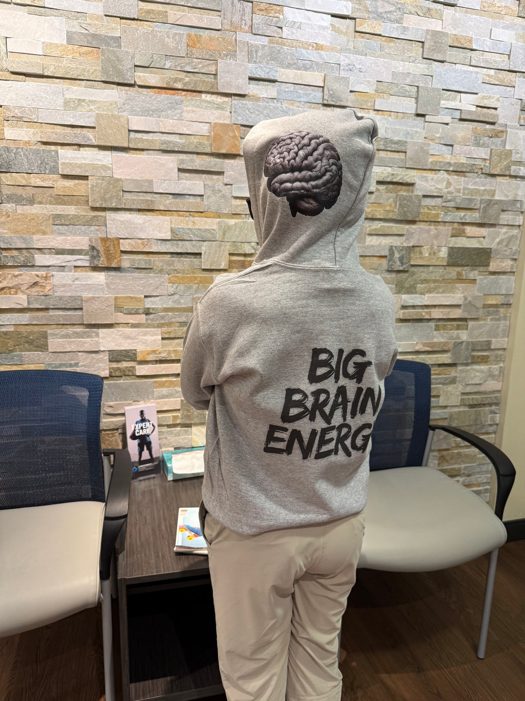 Child model wearing a light gray hoodie that says "BIG BRAIN ENERGY" in bold black capital letters on the back. The hood is up and shows the brain design on the left side.