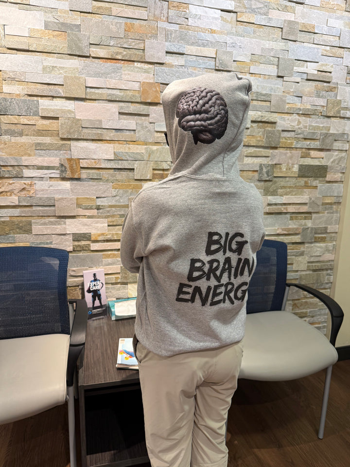 Child model wearing a light gray hoodie that says "BIG BRAIN ENERGY" in bold black capital letters on the back. The hood is up and shows the brain design on the left side.