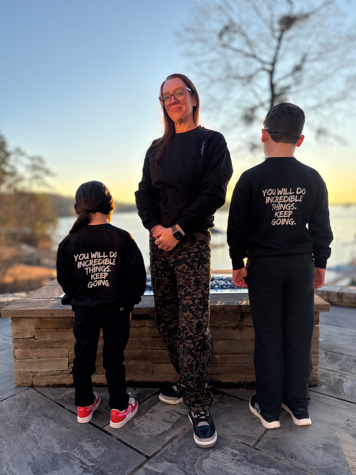 Dr. Betsy Grunch (Ladyspinedoc) facing forward wearing the black Incredible Sweatshirt. Youth models on either side of Dr. Grunch are facing backward, wearing the black youth versions of the Incredible Sweatshirt and showing the bold white brushstroke message: "YOU WILL DO INCREDIBLE THINGS. KEEP GOING." on the back.