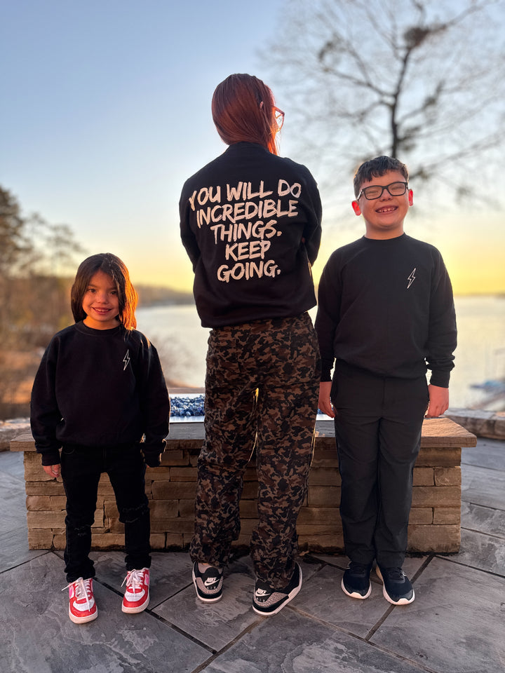 Dr. Betsy Grunch (Ladyspinedoc) facing backward wearing the black Incredible Sweatshirt showing the bold white brushstroke message: "YOU WILL DO INCREDIBLE THINGS. KEEP GOING." on the back. Youth models on either side of Dr. Grunch are facing forward, wearing the black youth versions of the Incredible Sweatshirt.