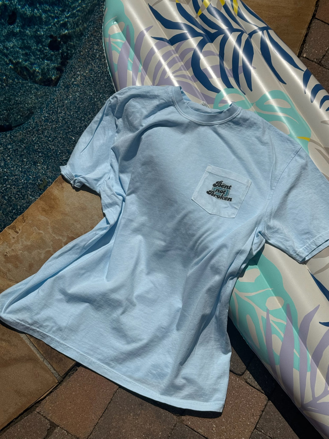 Light blue  Bent, Not Broken Scoliosis Awareness Tee - Blue Cotton T-Shirt, showing the pocket with the Bent not Broken catchphrase on a tropical-themed inflatable pool float.