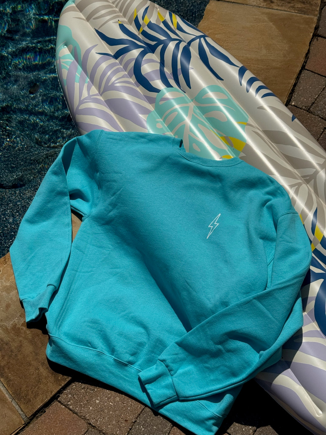 Teal sweatshirt on a tiled floor with a pool and inflatable raft in the background