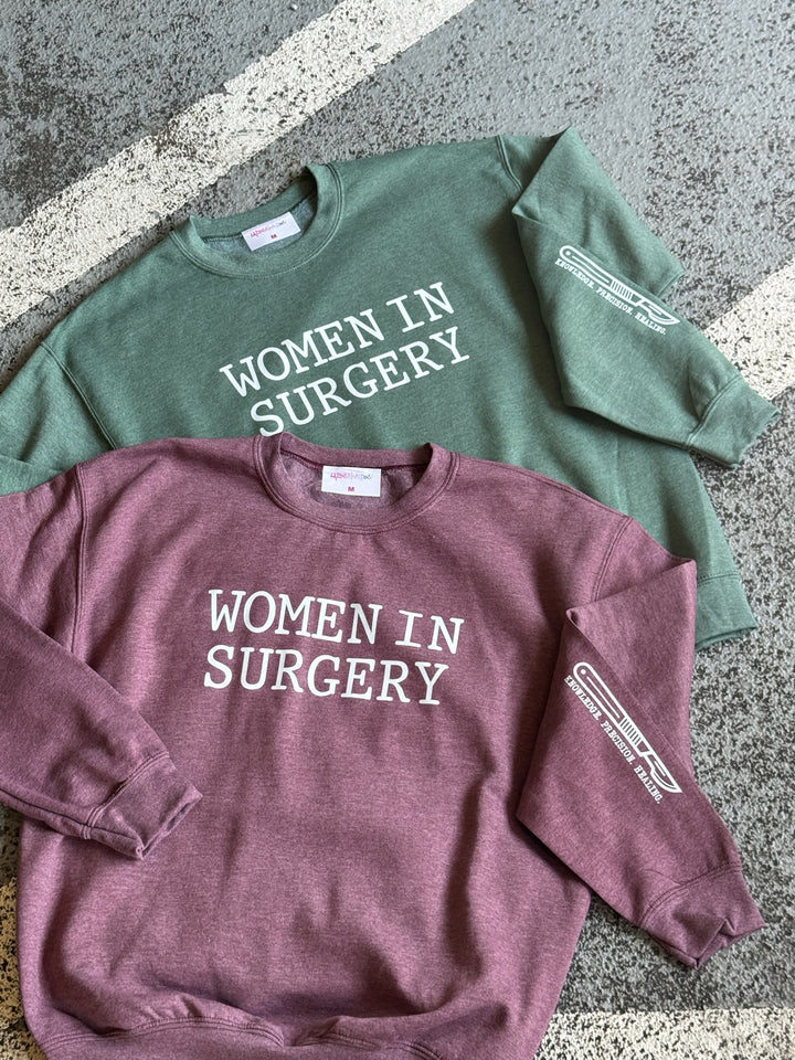 Flat lay of two 'Women in Surgery' crewneck sweatshirts on concrete, arranged with the mauve one on top of the green one. Both have a white scalpel graphic and 'Knowledge Precision Healing' printed on the sleeve.