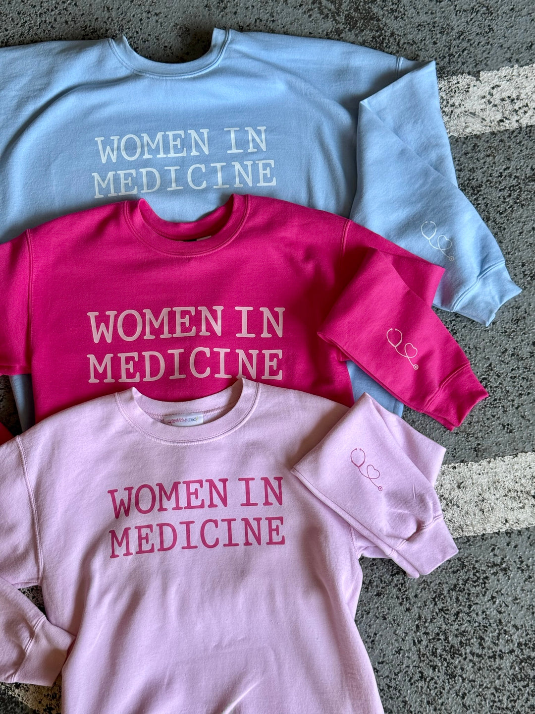 Light Pink, Light Blue, and Hot Pink Women in Medicine Crewneck Sweatshirts showing the Stethoscope Design on left sleeve.